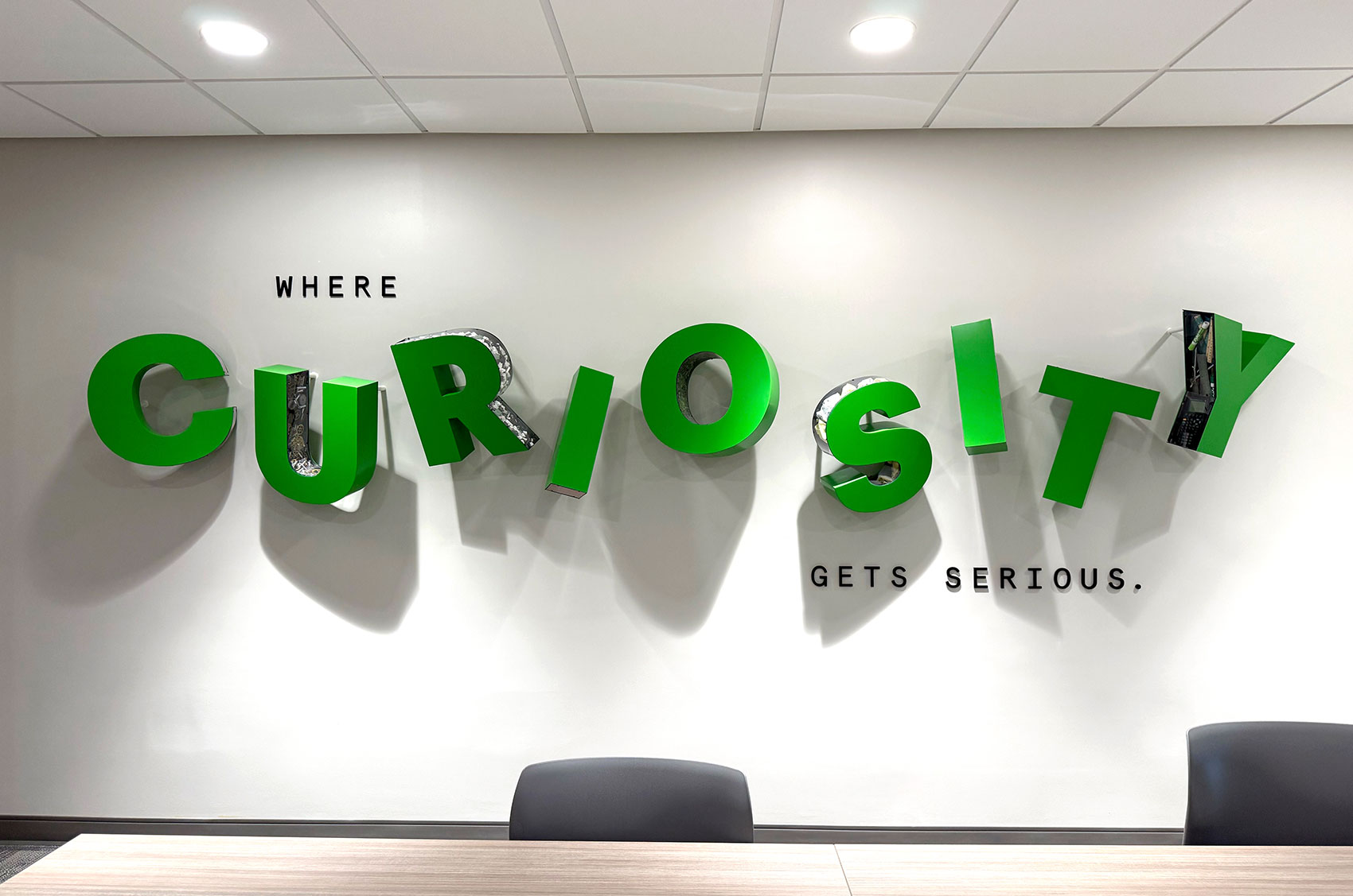 3D green letters spell CURIOSITY on a wall with Where Curiosity Gets Serious slogan. Modern office decor.